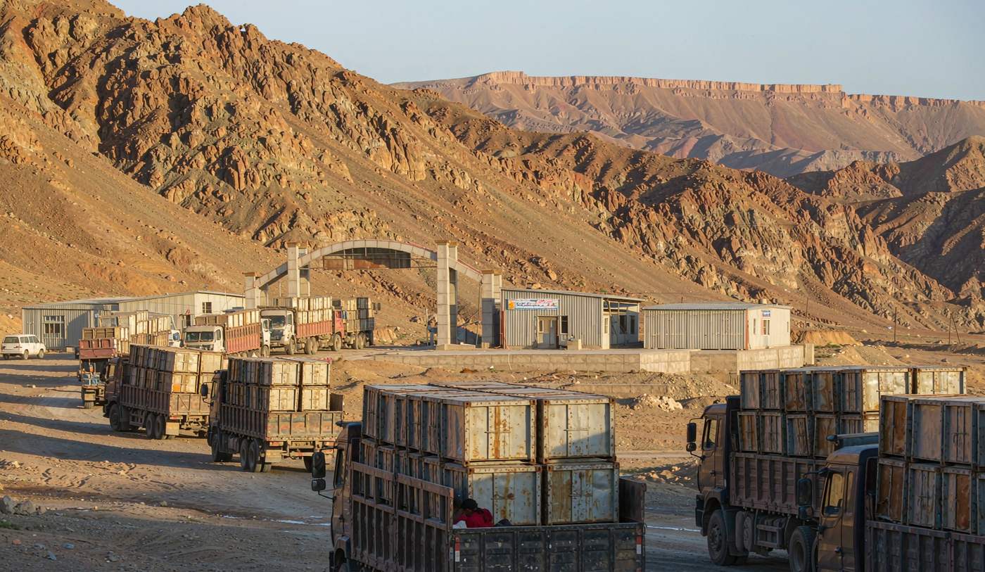 A mountainous Central Asian border crossing at dawn with a line of heavy cargo trucks and shipping containers waiting on a dusty road, a border gate and customs buildings ahead, rocky arid mountains behind