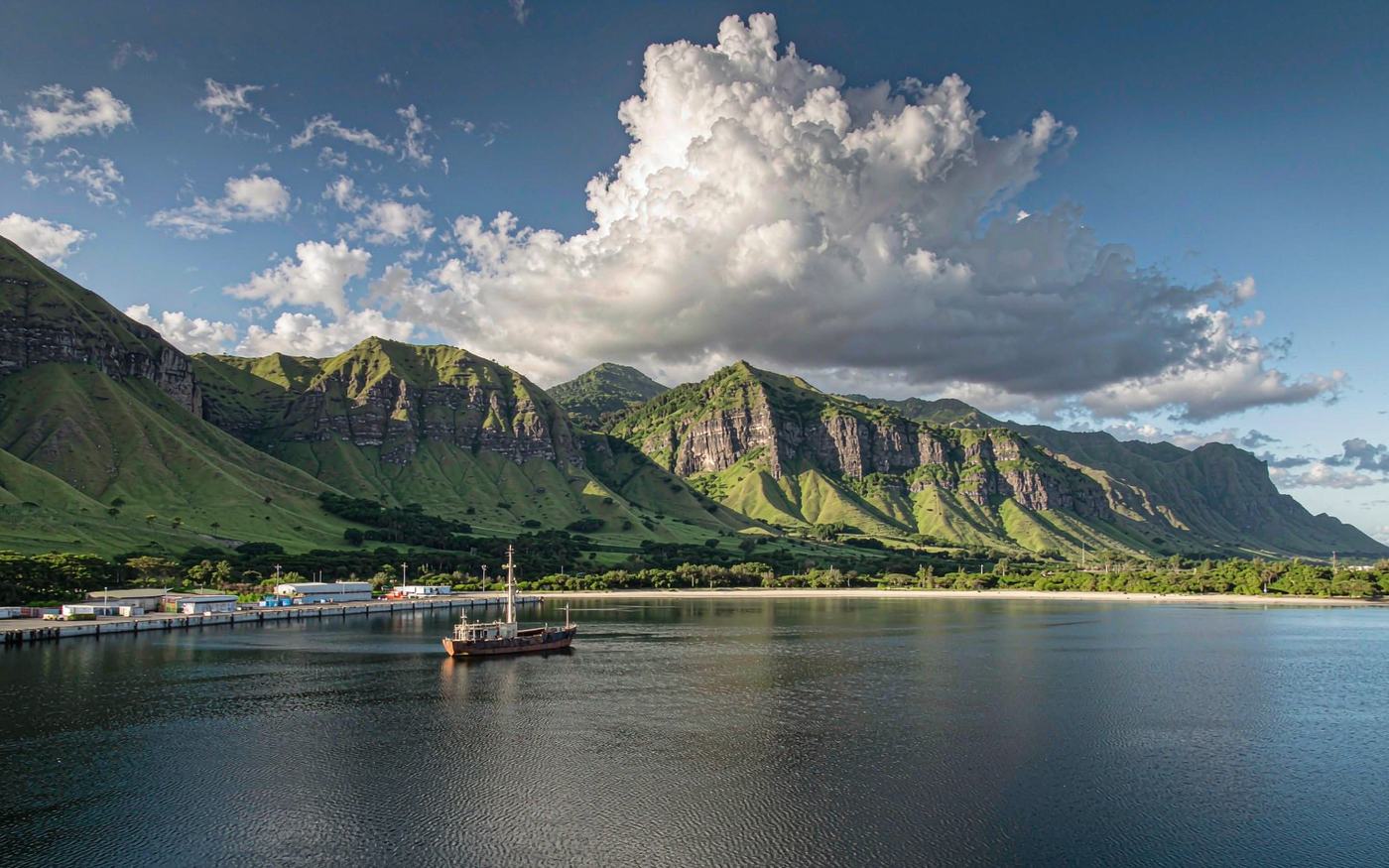 Photograph of a small tropical South Pacific harbour ringed by steep lush green volcanic mountains, calm dark blue water, a mid-sized cargo ship at anchor, low industrial wharf buildings and stacked containers
