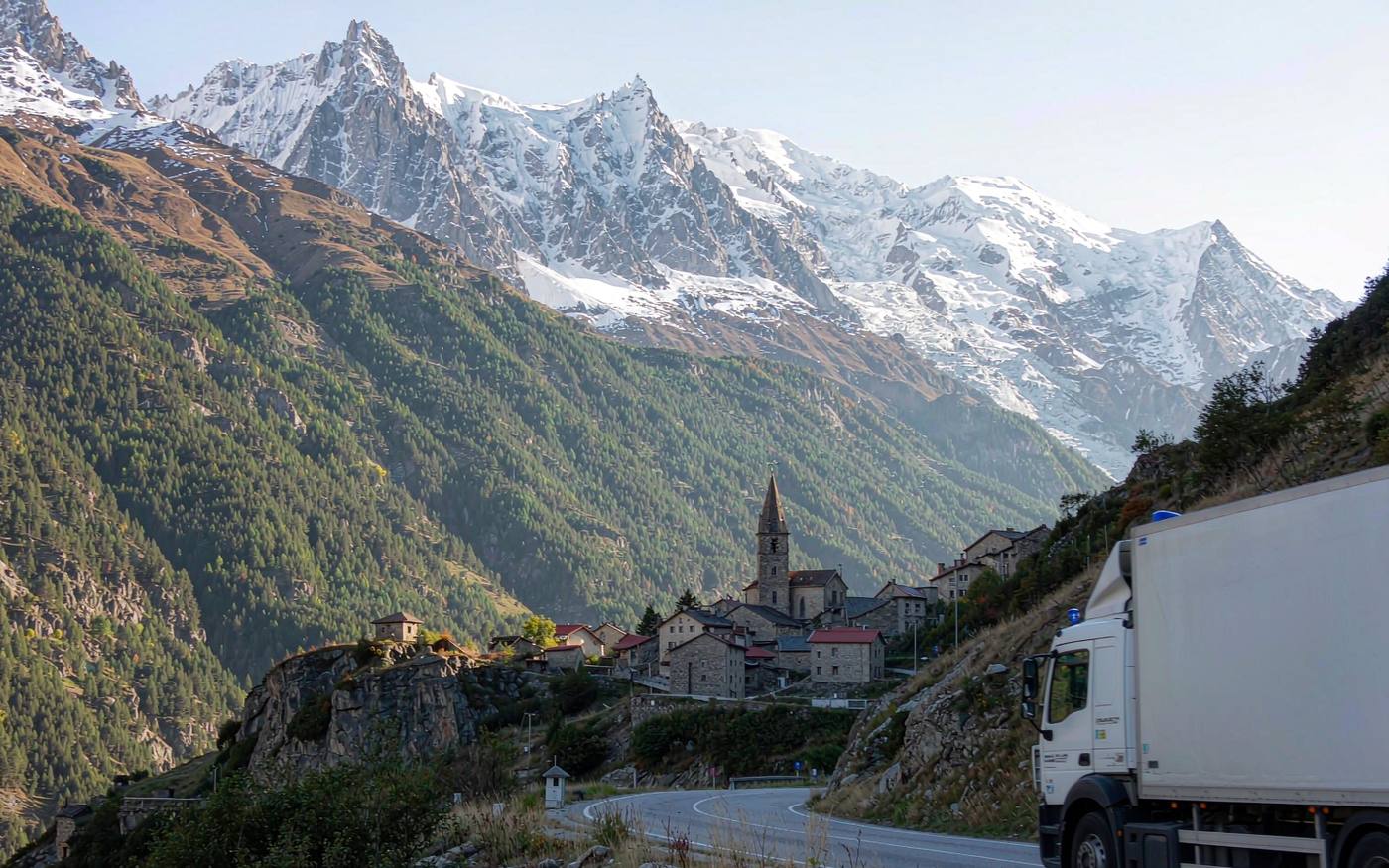 Delivery truck on a winding mountain road in the Pyrenees approaching a valley town with snow-capped peaks behind