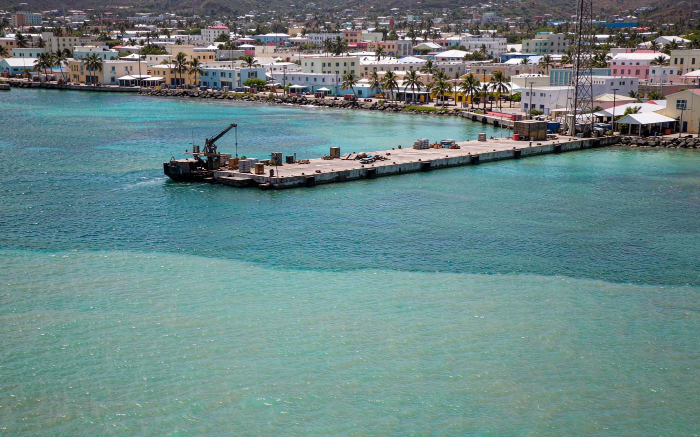 Wide photograph of a small Caribbean island harbour with turquoise shallow water, a modest concrete cargo jetty with a small landing craft unloading pallets and crates, low-rise white and pastel buildings and palm trees behind, bright midday light