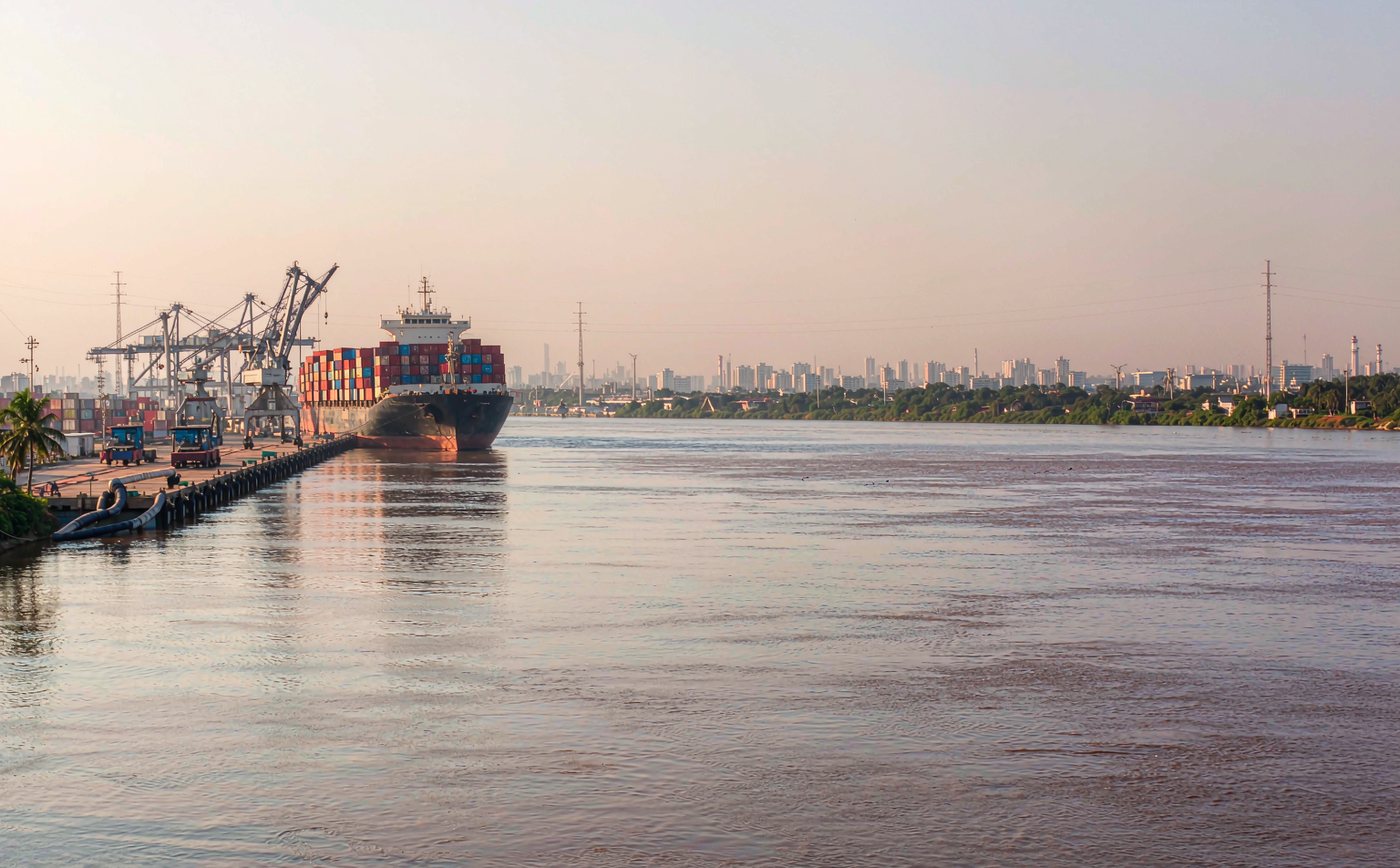 A South American river estuary container port in soft morning light with a container vessel at a long quay, gantry cranes, rows of stacked shipping containers and a low-rise port city skyline