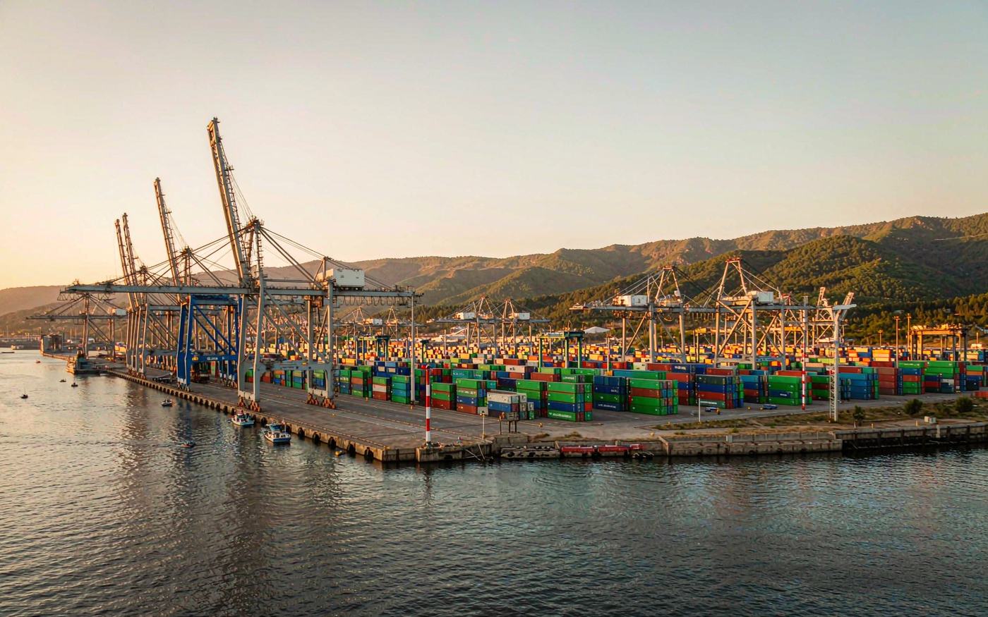 Photograph of a container port on the Black Sea in the Caucasus at golden hour with gantry cranes, stacked containers in green blue and red and green hills behind