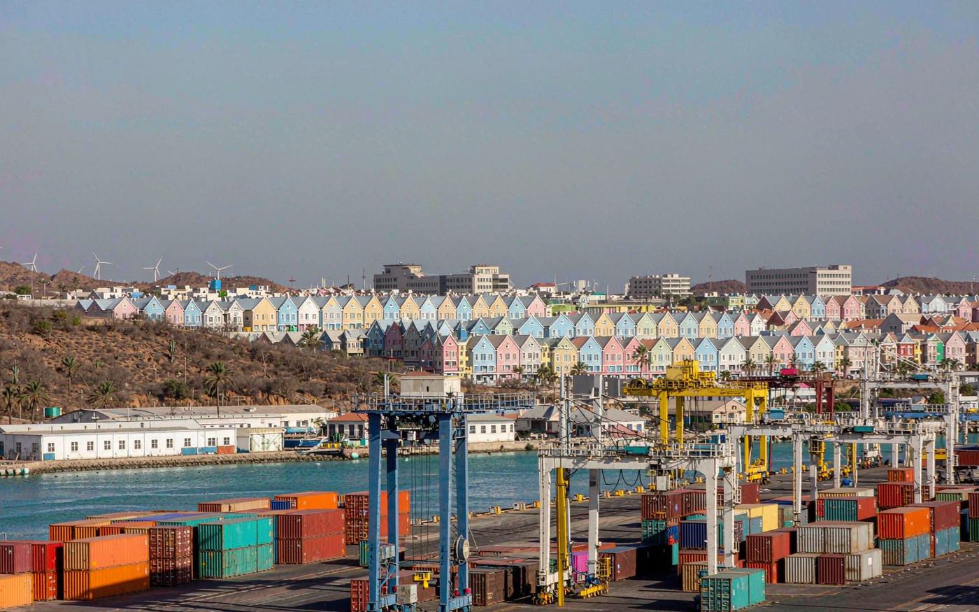 Wide photograph of a modern Caribbean container terminal with a ship-to-shore gantry crane and stacked containers in a paved yard, arid cactus-covered hills and pastel Dutch colonial gabled buildings of Oranjestad in the distance under a bright blue sky, turquoise water at the edge