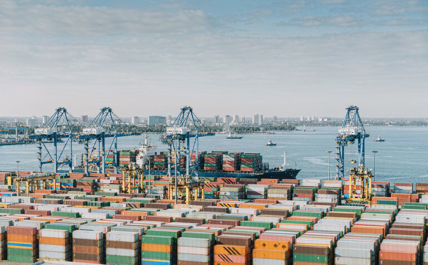 A large Australian container terminal in clear daylight, rows of stacked shipping containers, tall gantry cranes, a container vessel at the quay, calm harbour water and a distant city skyline