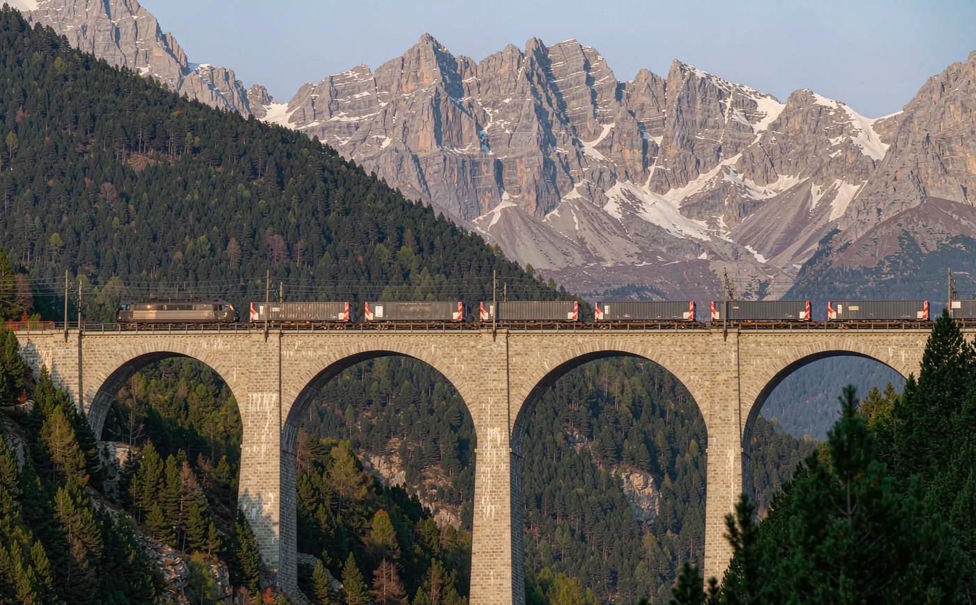 Freight train with intermodal container wagons crossing a high stone arch viaduct in the Austrian Alps, with forested slopes and snow-capped peaks behind