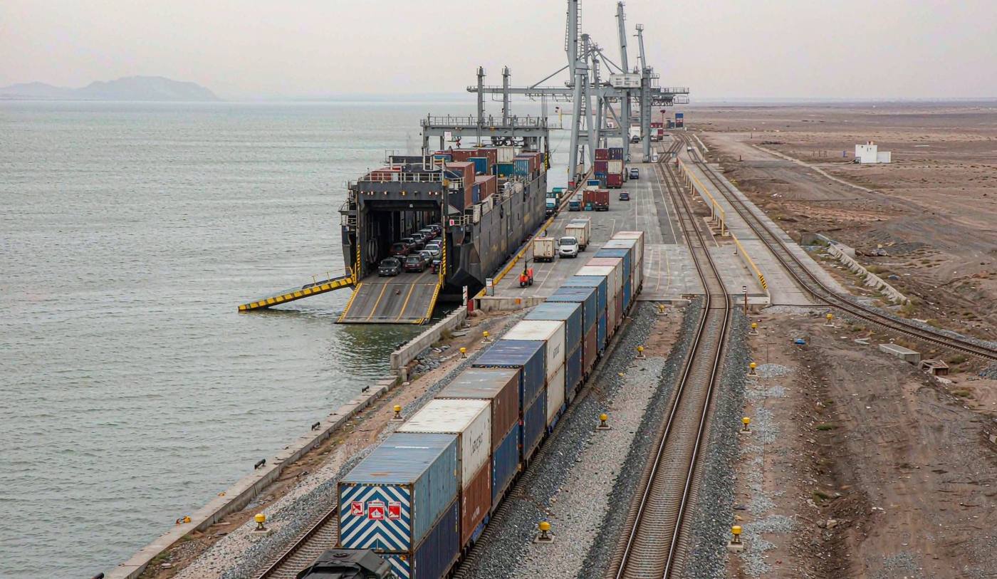 A Caspian Sea port rail terminal with intermodal freight trains beside a roll-on ferry ramp, stacked shipping containers and rail-mounted gantry cranes, calm grey-green sea and a flat semi-desert horizon behind under soft overcast light