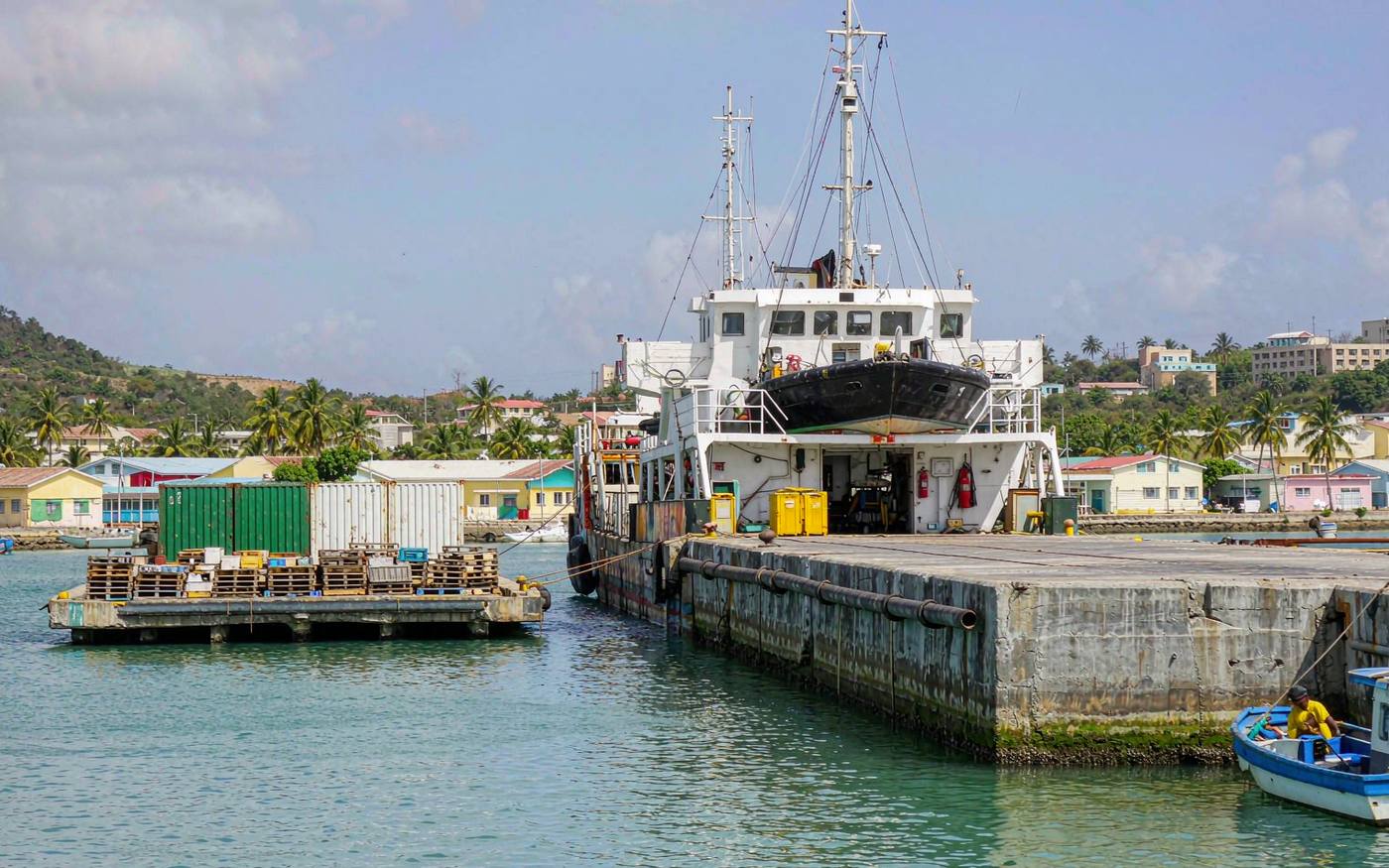 Caribbean island cargo quay with a small feeder ship at a concrete wharf, palletised freight and containers on the apron