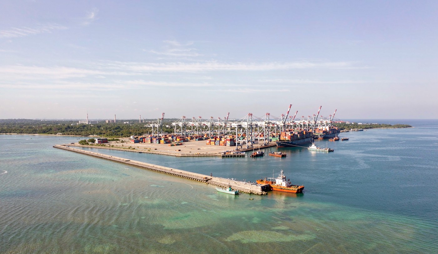 Aerial view of a container port on a Gulf island with gantry cranes over stacked shipping containers, a container vessel at berth, and a long low causeway crossing shallow turquoise water towards a distant shoreline under bright midday sun