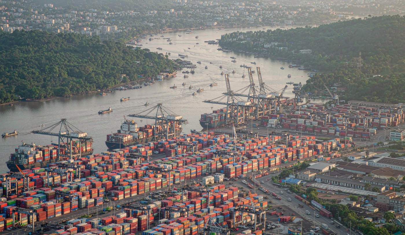 Aerial photograph of Chittagong port in Bangladesh on the Karnaphuli river with a dense container yard packed with stacked shipping containers, gantry cranes along the quay, two container vessels berthed, small cargo boats on the river, green hills and dense city rooftops under humid late afternoon light