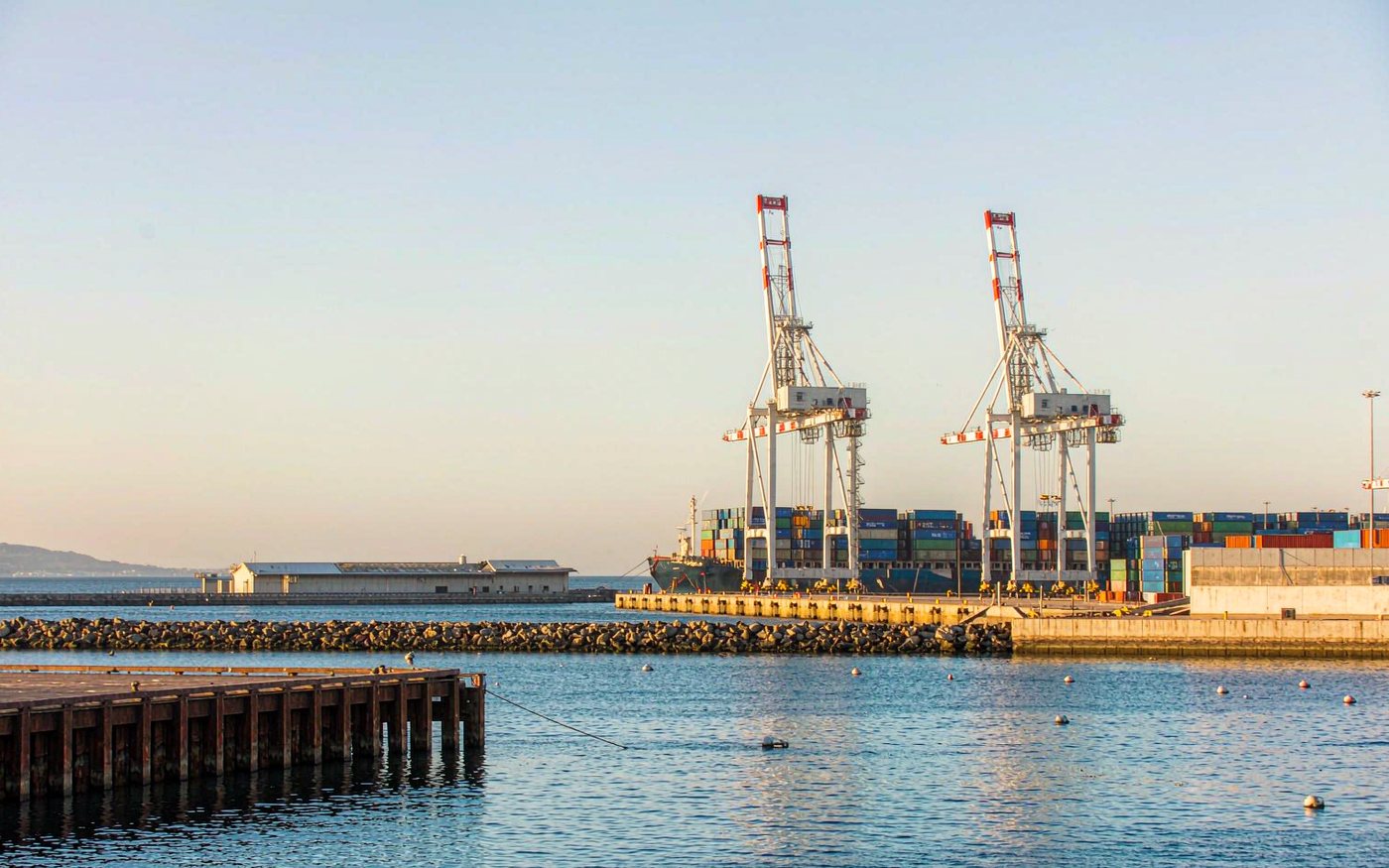 Caribbean container port with two ship-to-shore gantry cranes, stacked containers, a warehouse shed and a breakwater