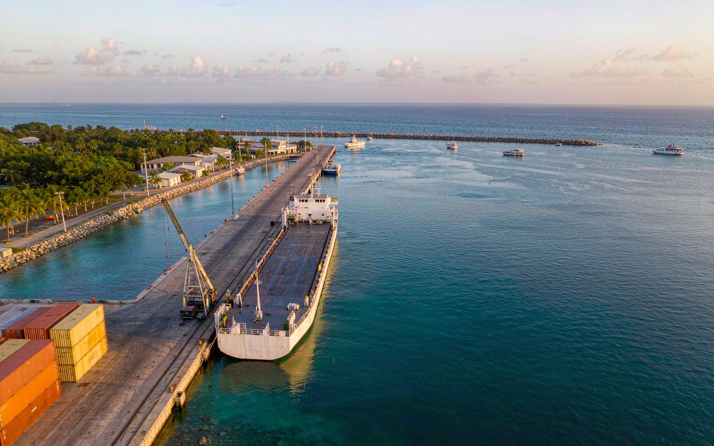 Documentary photograph of the Port of Belize City on the Caribbean coast of Central America, a small container terminal with stacked shipping containers and a mobile harbour crane, a cargo vessel at berth, palm trees and low tropical buildings along the shore, calm turquoise water under warm hazy afternoon light