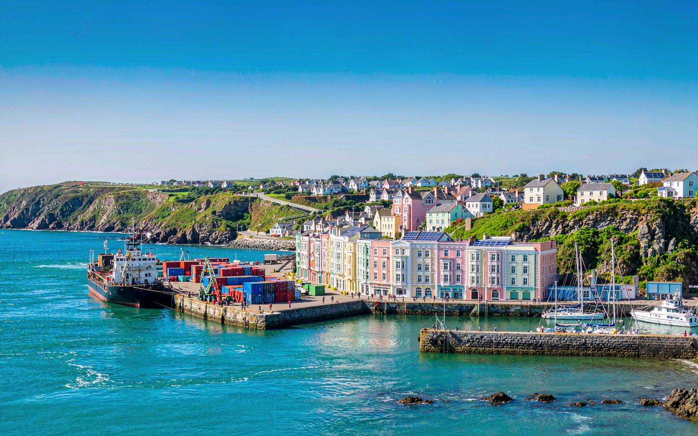 Elevated photograph of a small North Atlantic island harbour with calm turquoise water, pastel colonial harbourfront buildings in pink and coral tones along the quay, a small cargo ship moored at the wharf, stacked shipping containers and moored sailboats