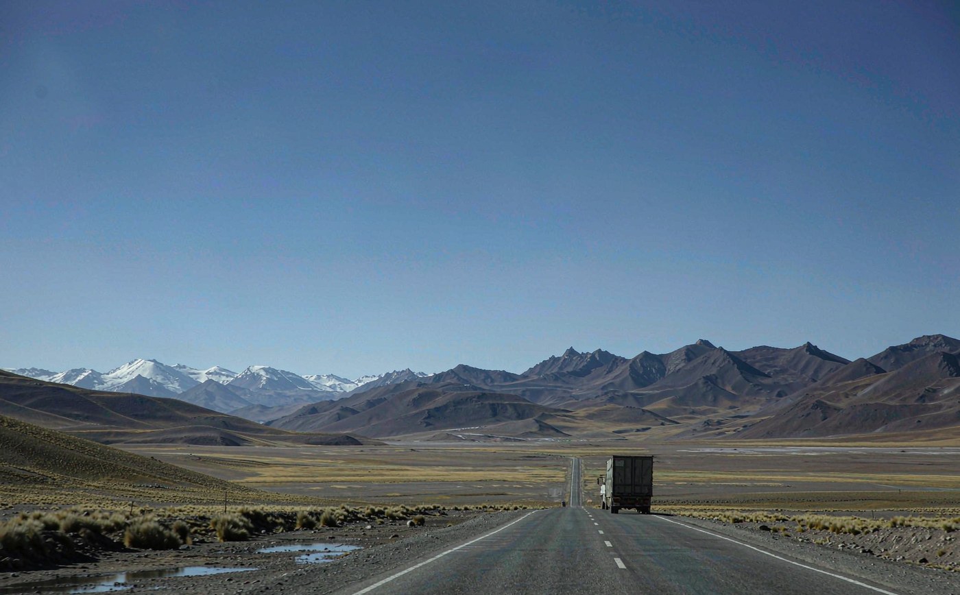 A container truck convoy on a high altitude Andean highway crossing a wide treeless altiplano plateau, with snow-capped peaks in the distance under a deep blue sky and dry golden grassland either side of the road