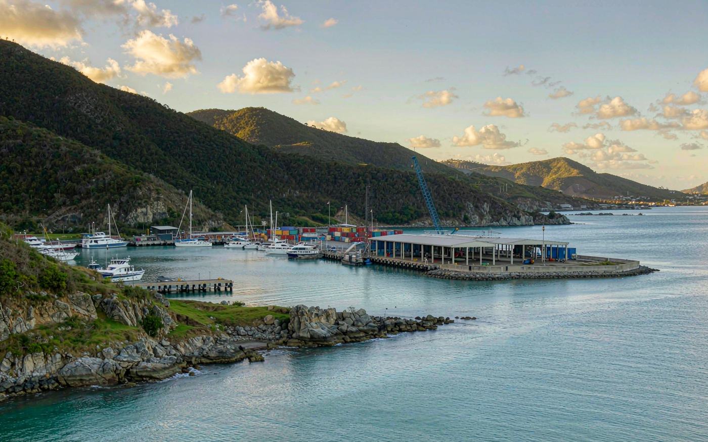Wide photograph of a Caribbean island harbour with turquoise water, a small working cargo quay with stacked containers and a mobile crane, low-rise white and pastel buildings and green hills behind, and sailing yachts at anchor offshore