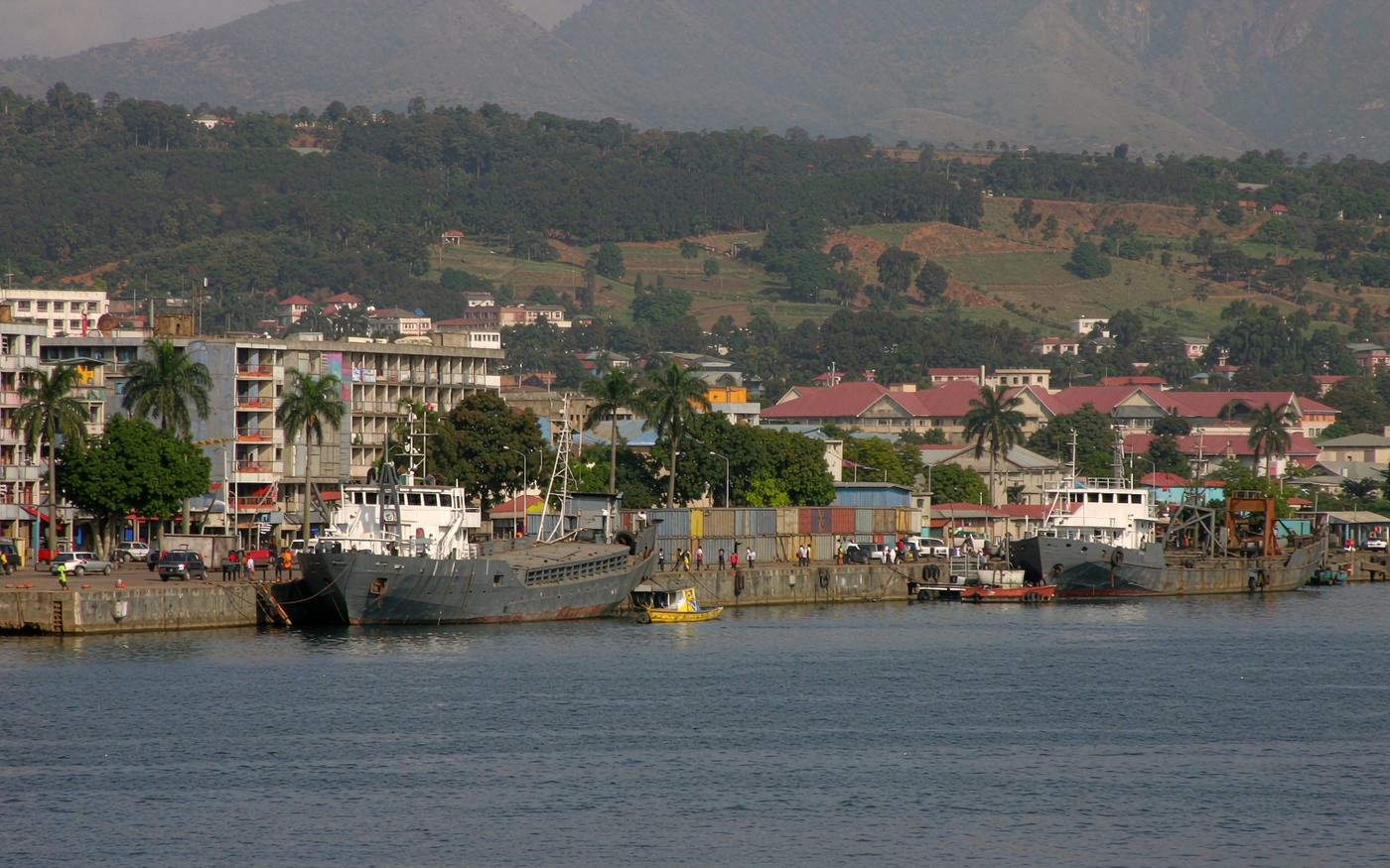 Lake port on Lake Tanganyika with a concrete quay and stacked containers, a lake cargo vessel moored alongside, cranes and low red-roofed buildings behind, palm trees along the shore and green Rift Valley hills rising beyond