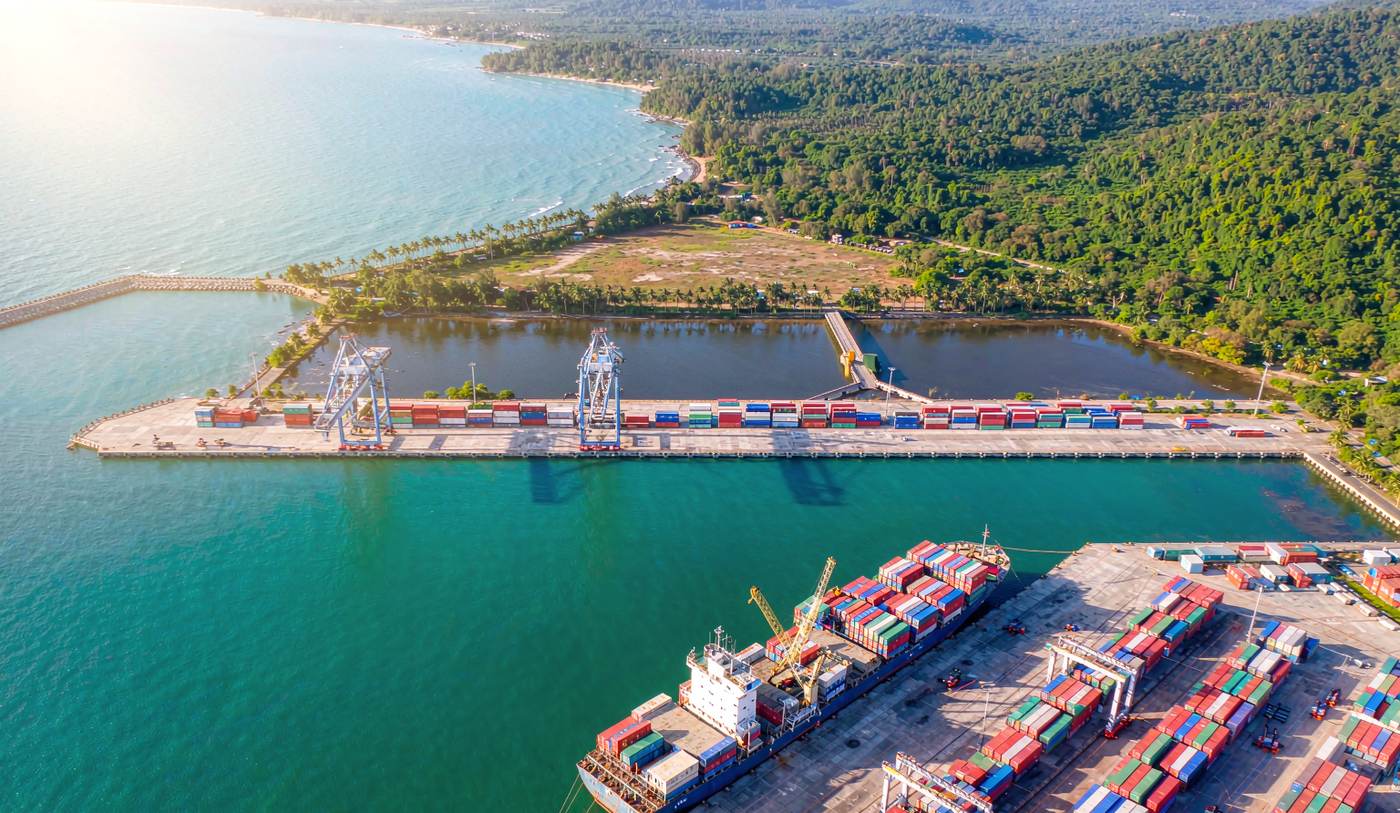 Aerial photograph of a container port on the Gulf of Thailand coast at Sihanoukville with a vessel at berth, gantry cranes over stacked shipping containers, a breakwater and calm turquoise water, palms and low green hills behind under midday light