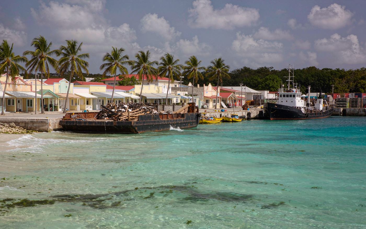 Photograph of a Caribbean island waterfront working port with extremely clear turquoise shallow water, a small cargo barge and a coaster at the quay, stacks of shipping containers on a modest terminal apron, palm trees and low pastel commercial buildings behind
