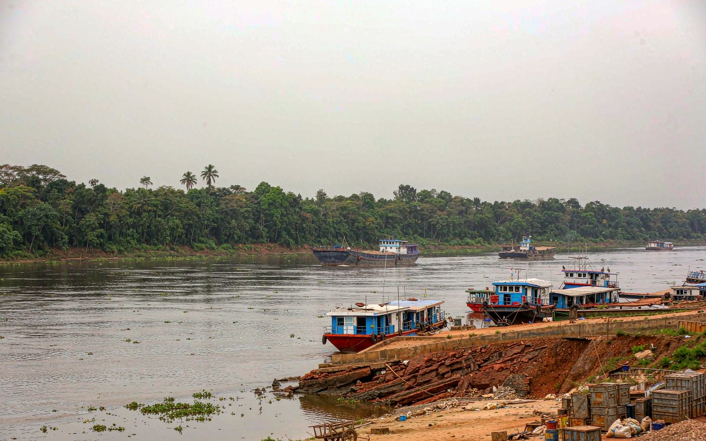 River port scene on a wide brown-green tropical river with barges and a small vessel moored at a simple concrete quay, cargo and containers stacked on red laterite soil, dense equatorial forest on the far bank under a humid overcast sky