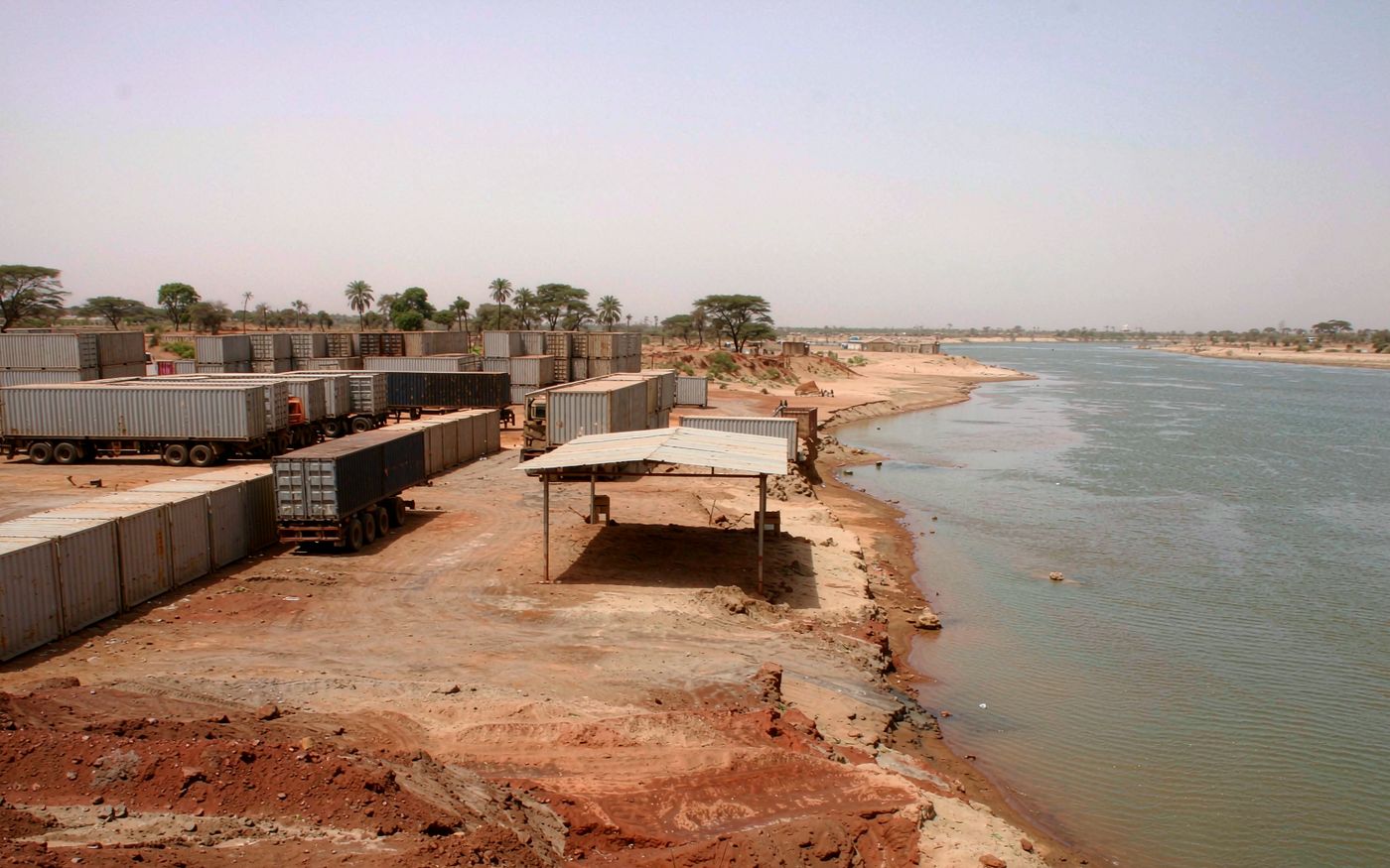 Inland logistics yard on the Sahel with container trailers and stacked containers on red-brown laterite earth, flat dry savanna with acacia trees, a wide shallow river with sandy banks in the distance and a hazy bright horizon
