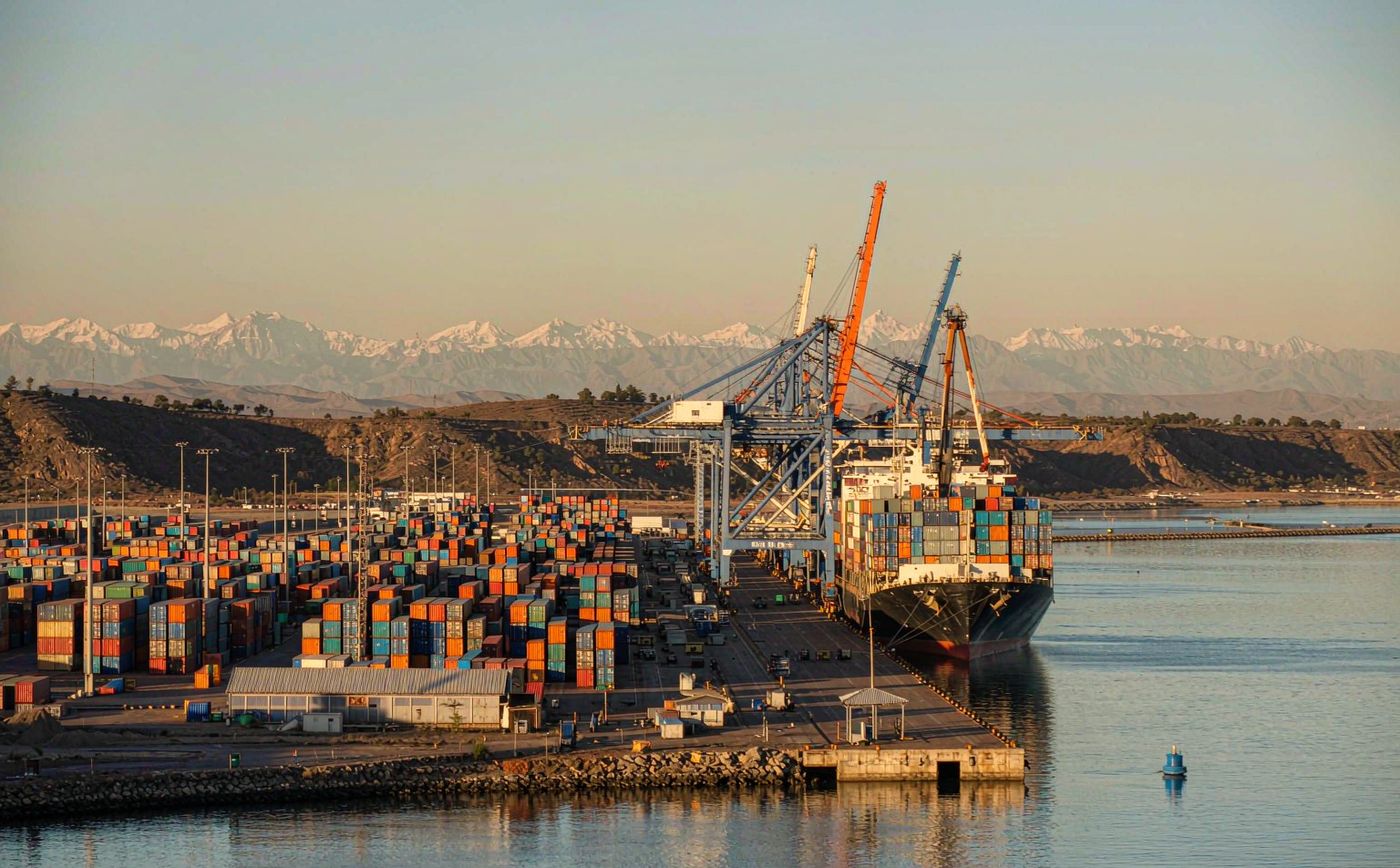 A Pacific container terminal in Chile at golden morning light with a container vessel at the quay, blue gantry cranes, stacked container yards, calm harbour water and distant snow-capped Andes on the horizon