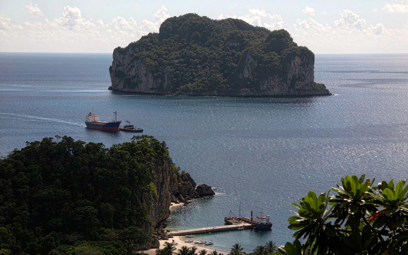 A cargo ship anchored off a steep green tropical island in the Indian Ocean with cargo being lifted onto a small wharf