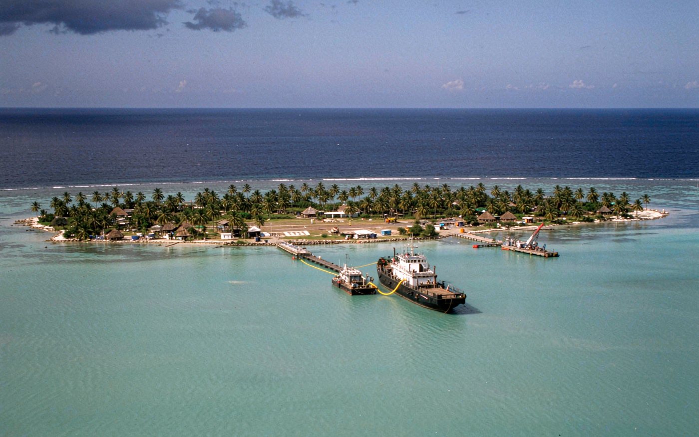 Indian Ocean coral atoll with a turquoise lagoon, coconut palms, a small airstrip and a supply vessel anchored offshore with a barge