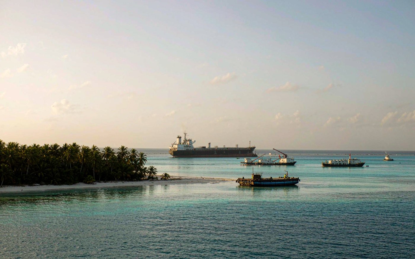 Indian Ocean coral atoll anchorage with cargo being lightered by barge from a ship standing off the reef, white sand beach and dense coconut palms ashore, clear turquoise lagoon