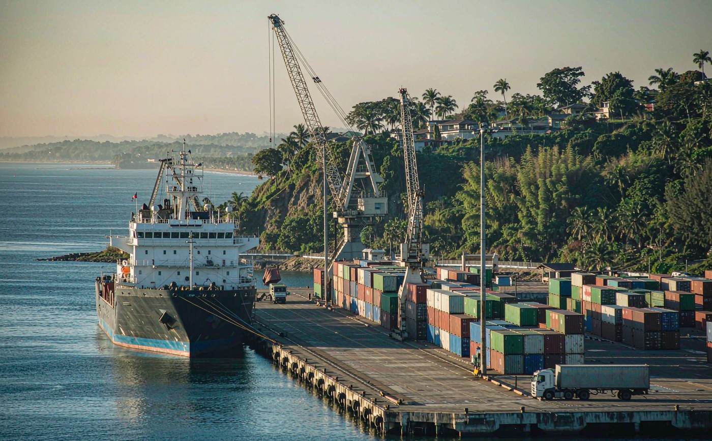 A Pacific container port in Colombia in bright morning light with a container vessel at the quay, gantry cranes, stacks of colourful shipping containers, calm estuary water and dense green tropical hills behind the terminal