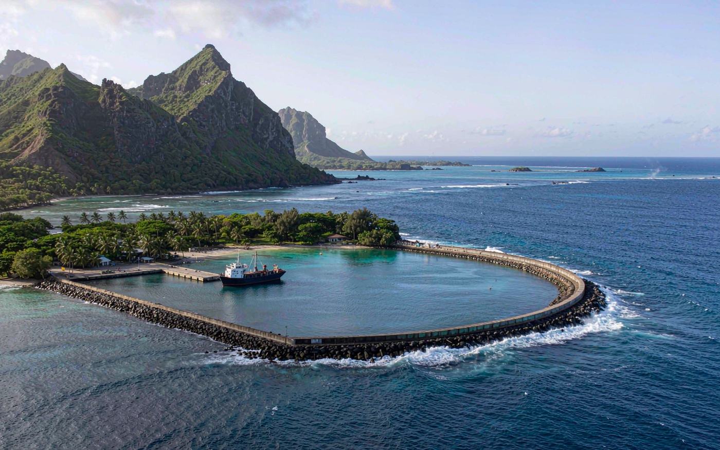 Wide photograph of a small South Pacific island harbour with a sheltered coral lagoon, a short concrete wharf with a small coastal cargo ship at berth, coconut palms and low green volcanic mountains behind, turquoise water and bright tropical light