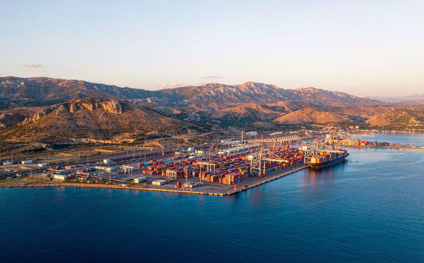 Aerial view of a container terminal on the Adriatic coast in soft morning light, gantry cranes over a modern quay, rows of shipping containers, a container ship alongside and karst mountains directly behind the port