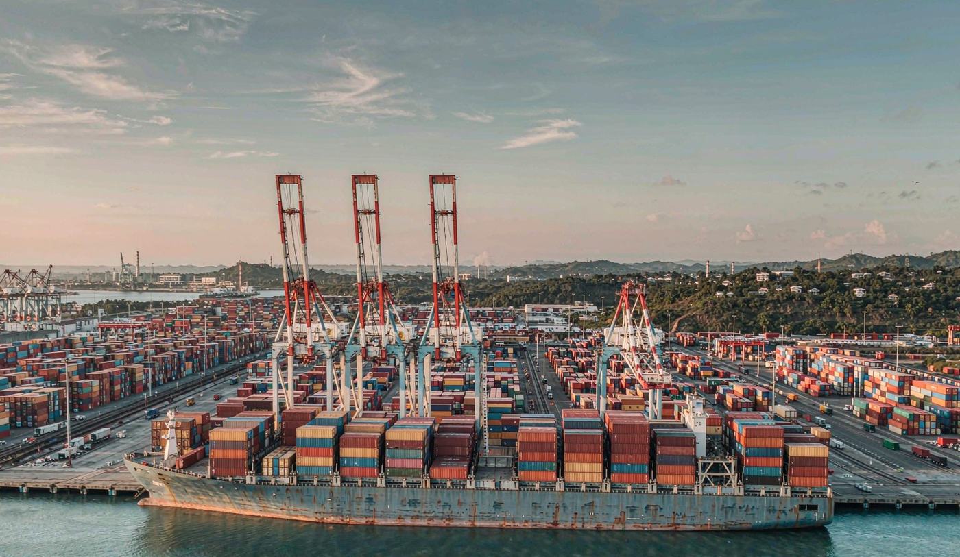 A large modern deep-water container terminal in the Caribbean with gantry cranes over rows of stacked shipping containers, a container vessel at berth, a wide paved yard and low tropical hills behind the port
