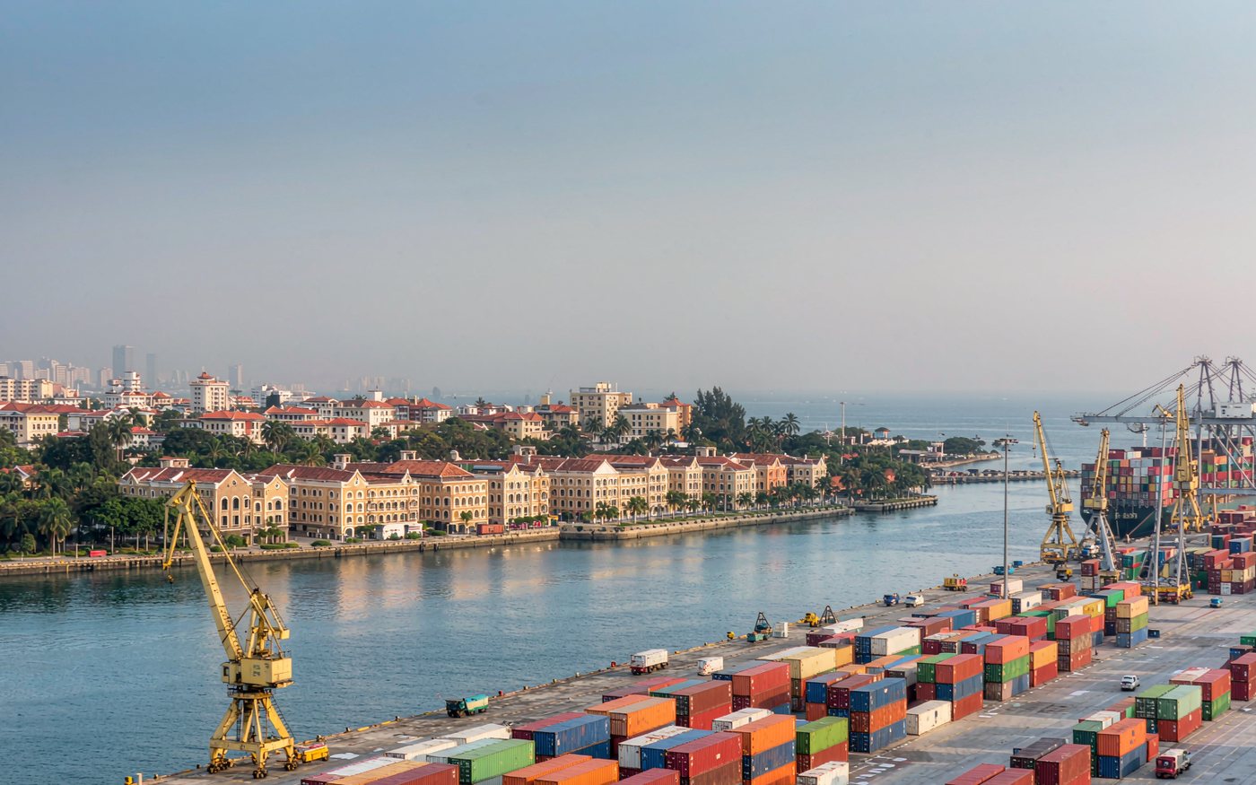Wide photograph of a deep Caribbean container harbour with a large gantry crane loading a container ship, rows of stacked containers in a paved yard, and pastel Dutch colonial waterfront buildings of Willemstad across the water under a bright sky