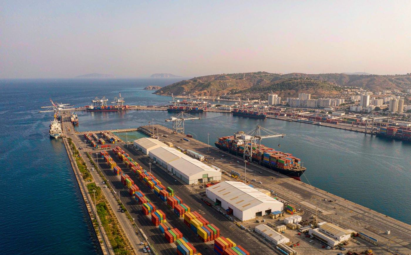 Aerial view of the container port of Limassol in bright eastern Mediterranean daylight, a long container quay with gantry cranes, dense rows of stacked shipping containers, a container vessel berthed and dry pale hills with a low city skyline behind