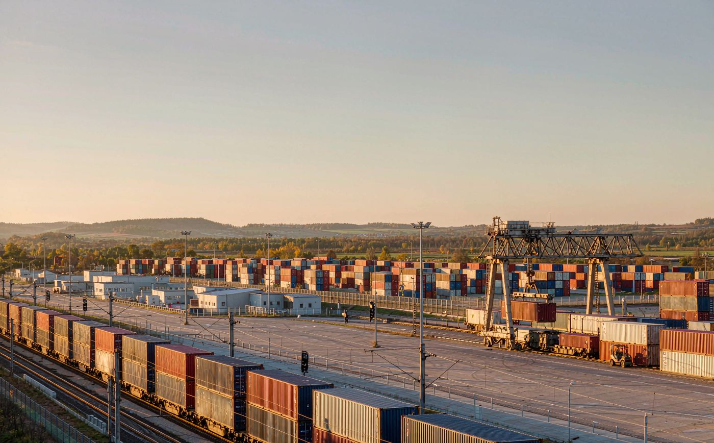 Inland rail container terminal at golden hour, intermodal wagons loaded with shipping containers, a rail-mounted gantry crane lifting a container and low forested hills behind