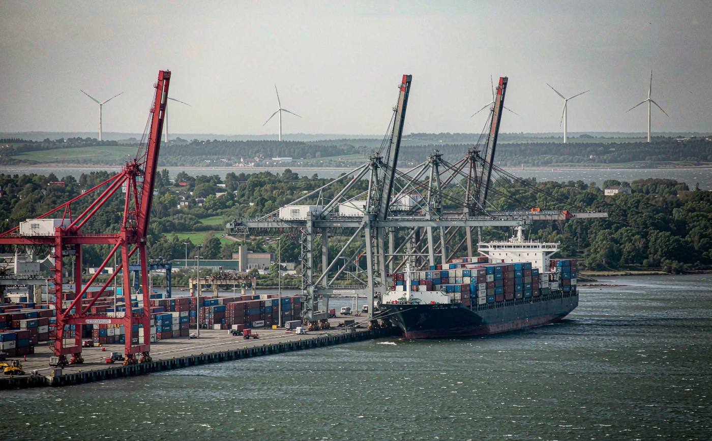 Wide view of a Danish container port with gantry cranes at the quay, rows of stacked containers in the yard, a container ship at berth and offshore wind turbines on the horizon