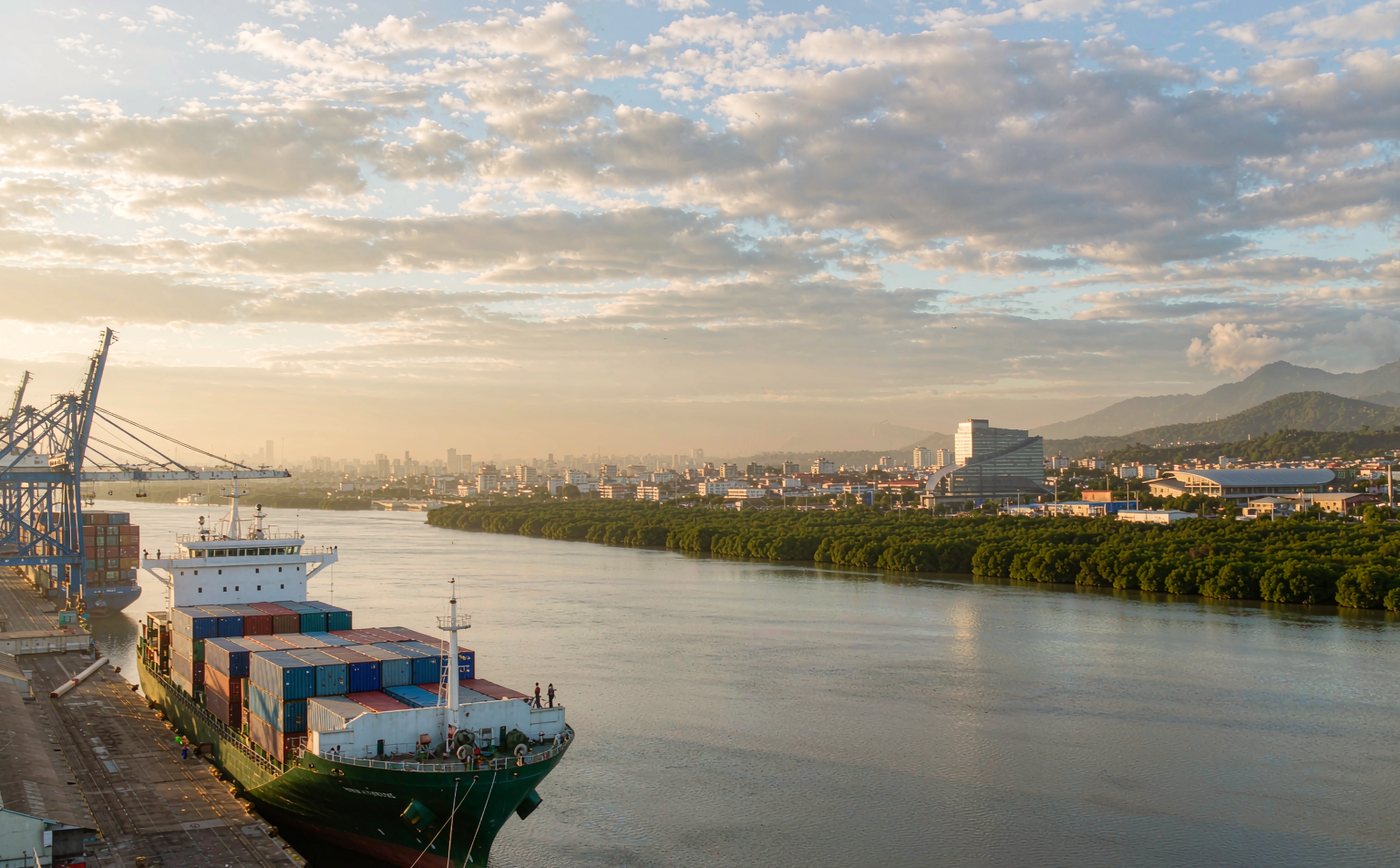 A container terminal on a broad tropical river estuary on Ecuador's Pacific coast in warm late-morning light, with a container ship at the quay, gantry cranes, stacked containers, mangrove vegetation, a low-rise port city skyline and distant green Andean foothills