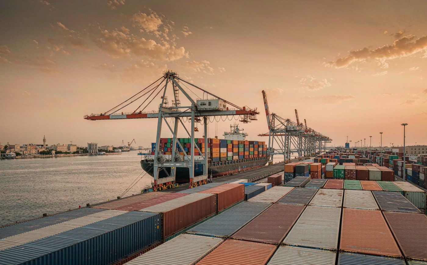 A Mediterranean container port at golden hour with gantry cranes loading a container vessel, rows of stacked shipping containers and a distant port skyline with minarets