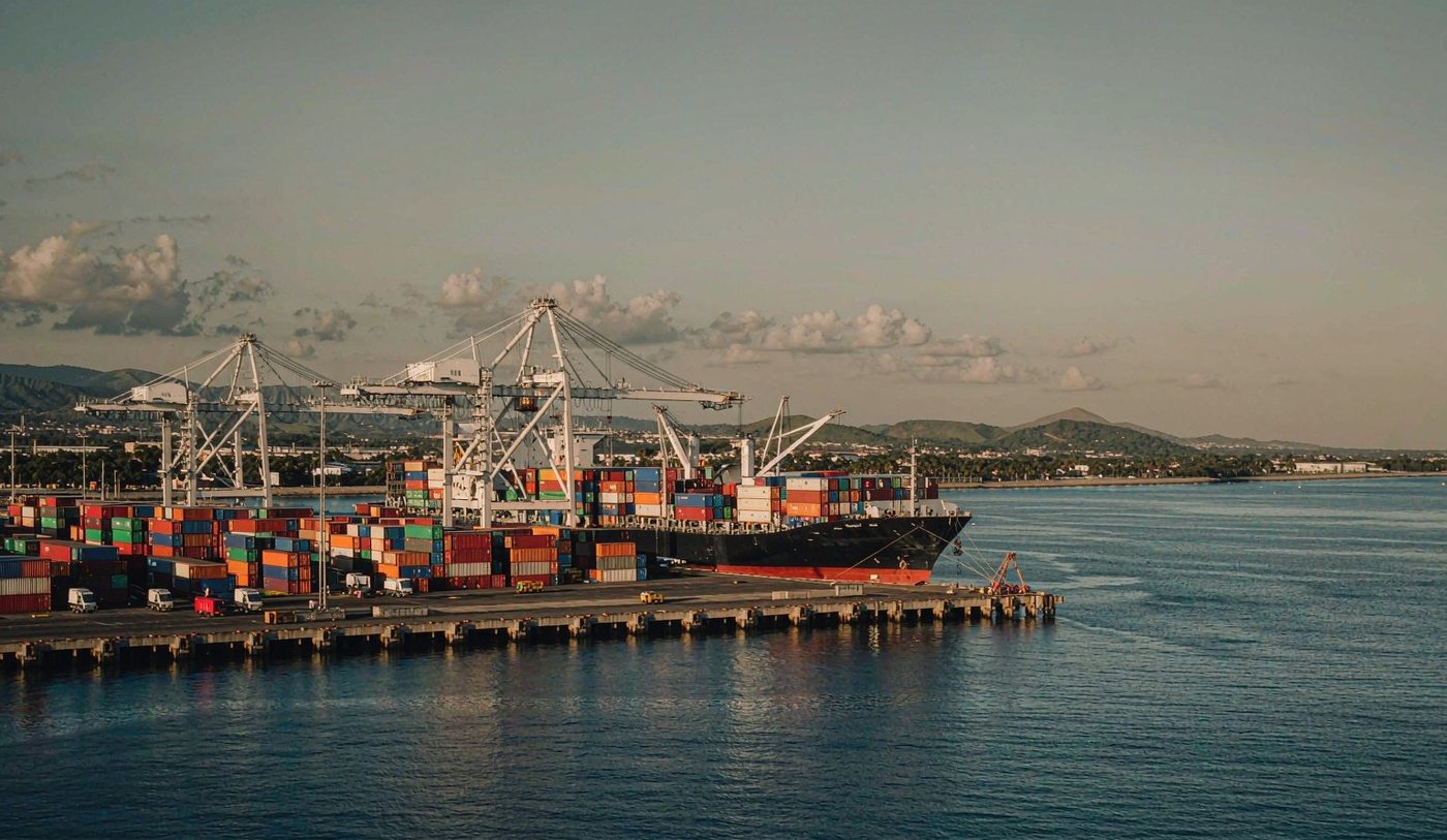 The container port of Acajutla on the Pacific coast of El Salvador with gantry cranes over stacked shipping containers, a container vessel at berth, calm harbour water and low tropical hills behind the coastline