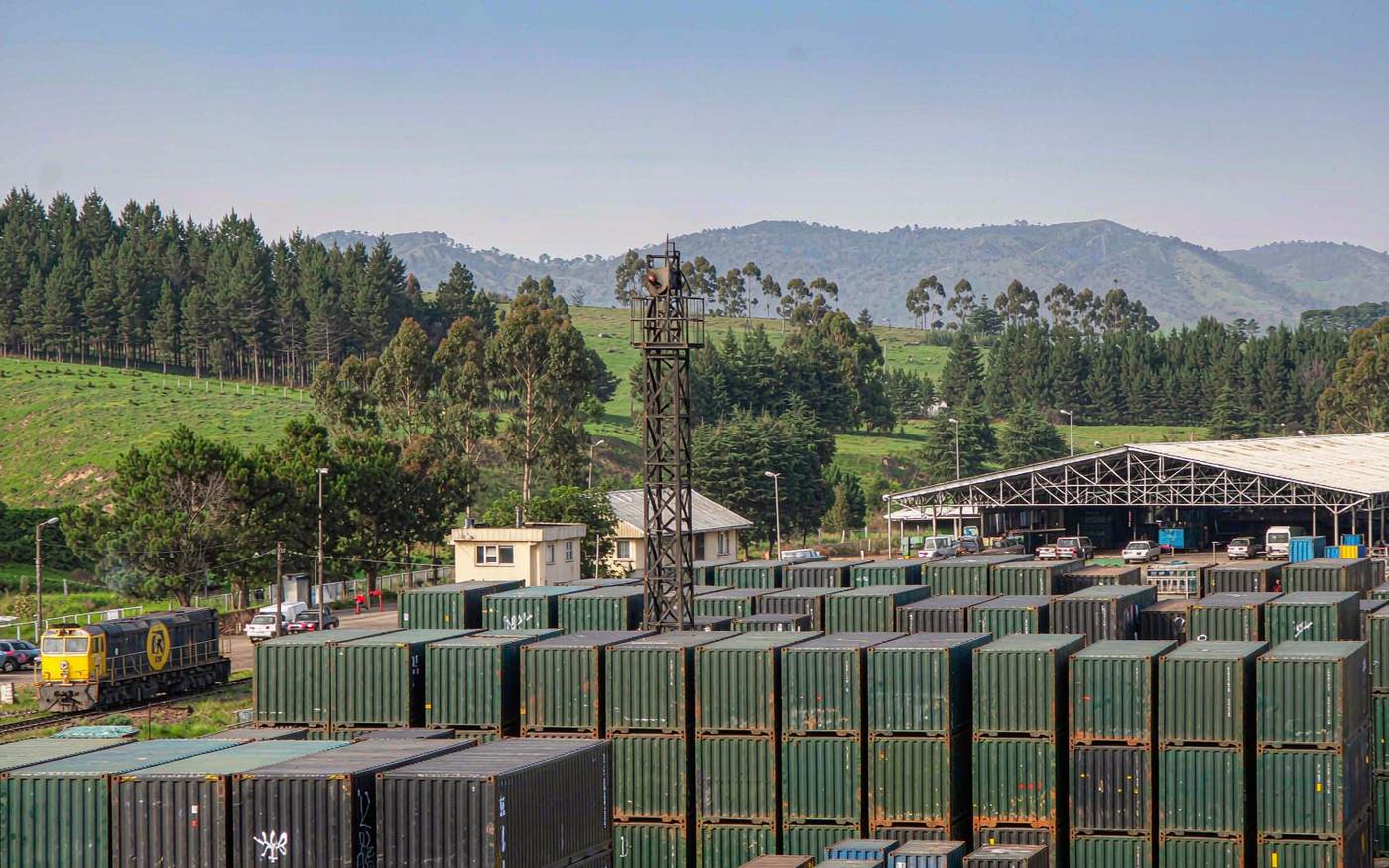 Small inland container depot with stacked shipping containers, a railway siding and a shunting locomotive, warehouse roofs, rolling green hills with pine and eucalyptus plantations behind and blue mountains on the horizon under clear high light