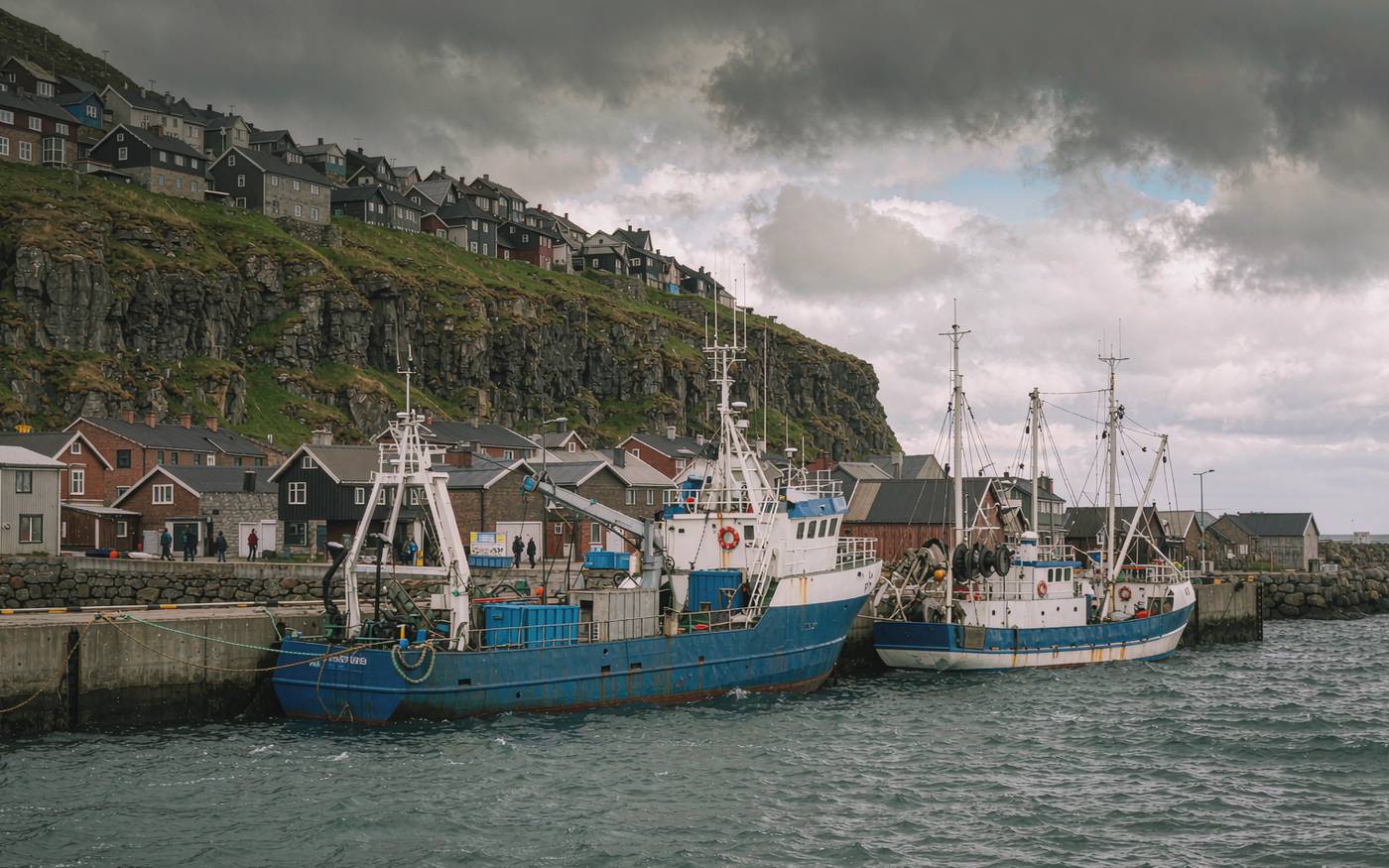 A cargo vessel and fishing boats at a quay in a North Atlantic harbour with grass cliffs behind
