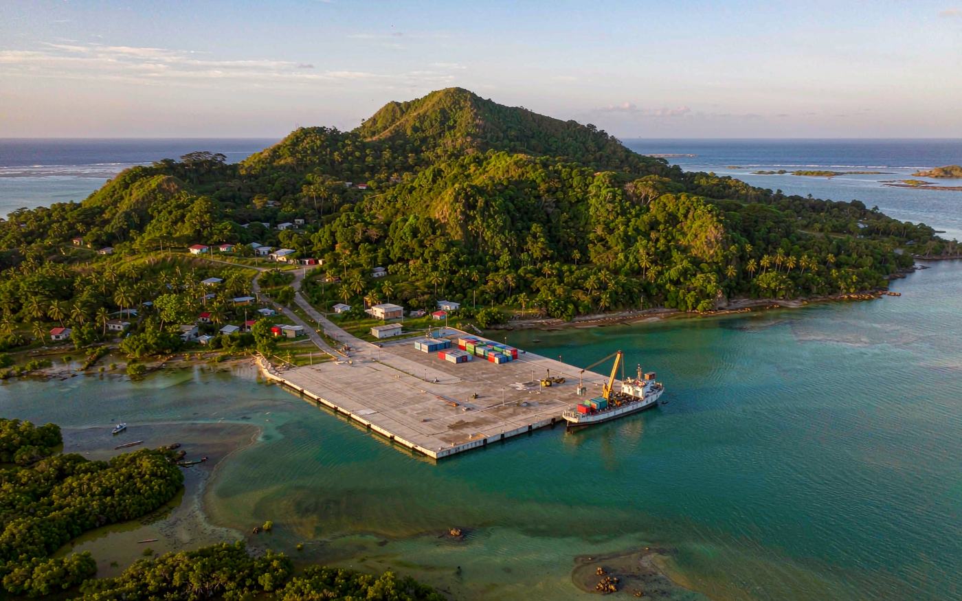 Small tropical island port with a concrete quay and stacked containers, a geared cargo ship alongside, dense green jungle hills behind, mangrove shoreline and turquoise lagoon in the foreground