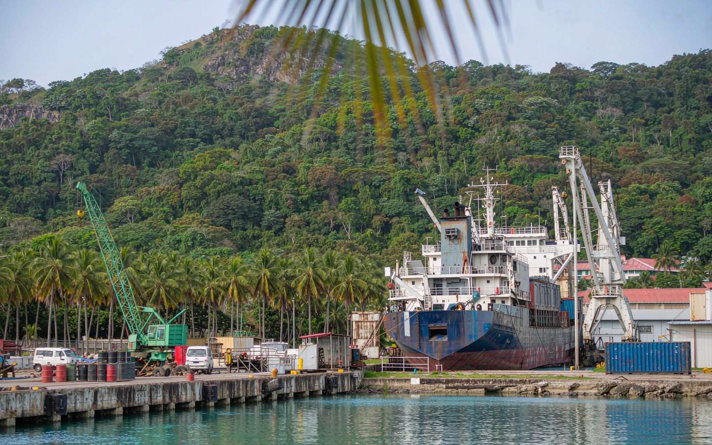 South Pacific container quay with a small ship-to-shore crane, stacked containers, a berthed feeder vessel and lush green hills behind