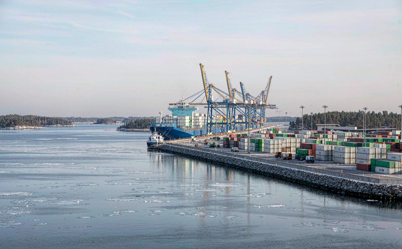 Nordic container terminal with gantry cranes along a granite quay, stacked shipping containers in the yard, a container vessel moored at the berth, calm Baltic water with light drift ice and low pine-covered islands on the horizon