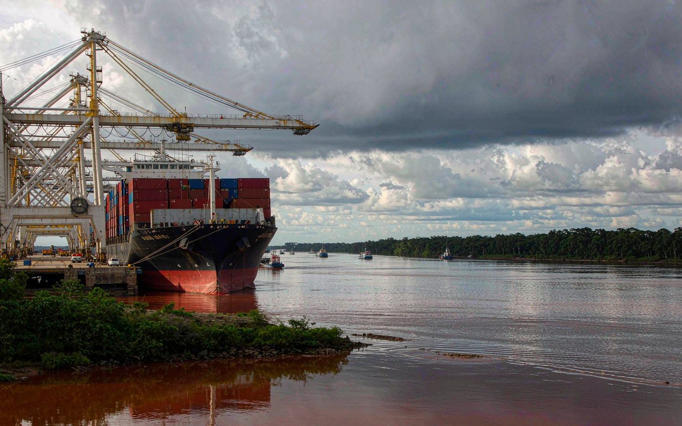 A container ship being worked by a gantry crane at a tropical river port with rainforest on the horizon