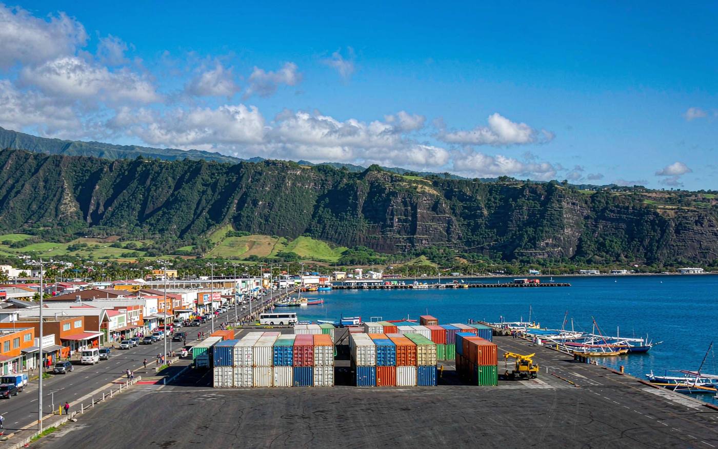 Wide photograph of Papeete harbour on Tahiti with a working cargo quay stacked with containers and a small mobile crane, colourful low-rise waterfront buildings, outrigger canoes in the water and a steep green mountainous island interior behind, bright tropical light