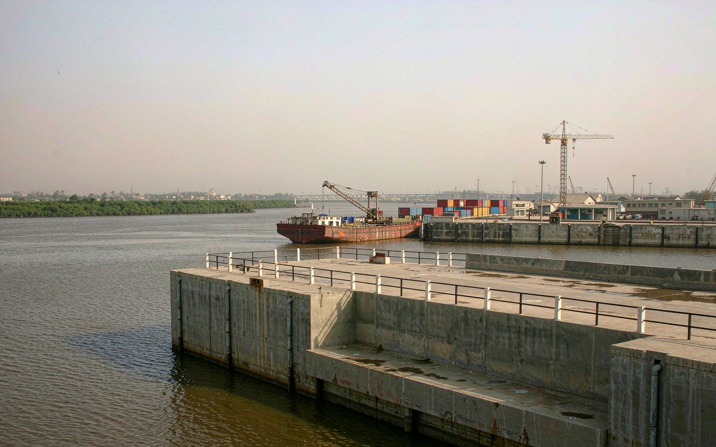 River port on a wide brown-green West African river with a concrete quay, stacked containers and a geared cargo ship alongside, low port buildings and cranes, mangrove vegetation on the far bank under warm hazy light