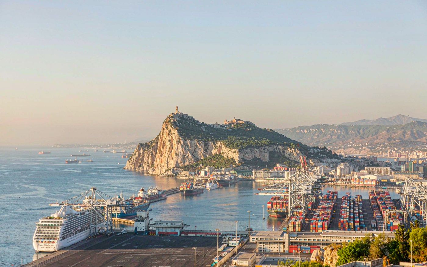Photograph of a Mediterranean port at the foot of a steep limestone rock peninsula, a container terminal with gantry cranes and stacked boxes, several cargo ships in the bay, calm blue strait water and hazy distant mainland hills