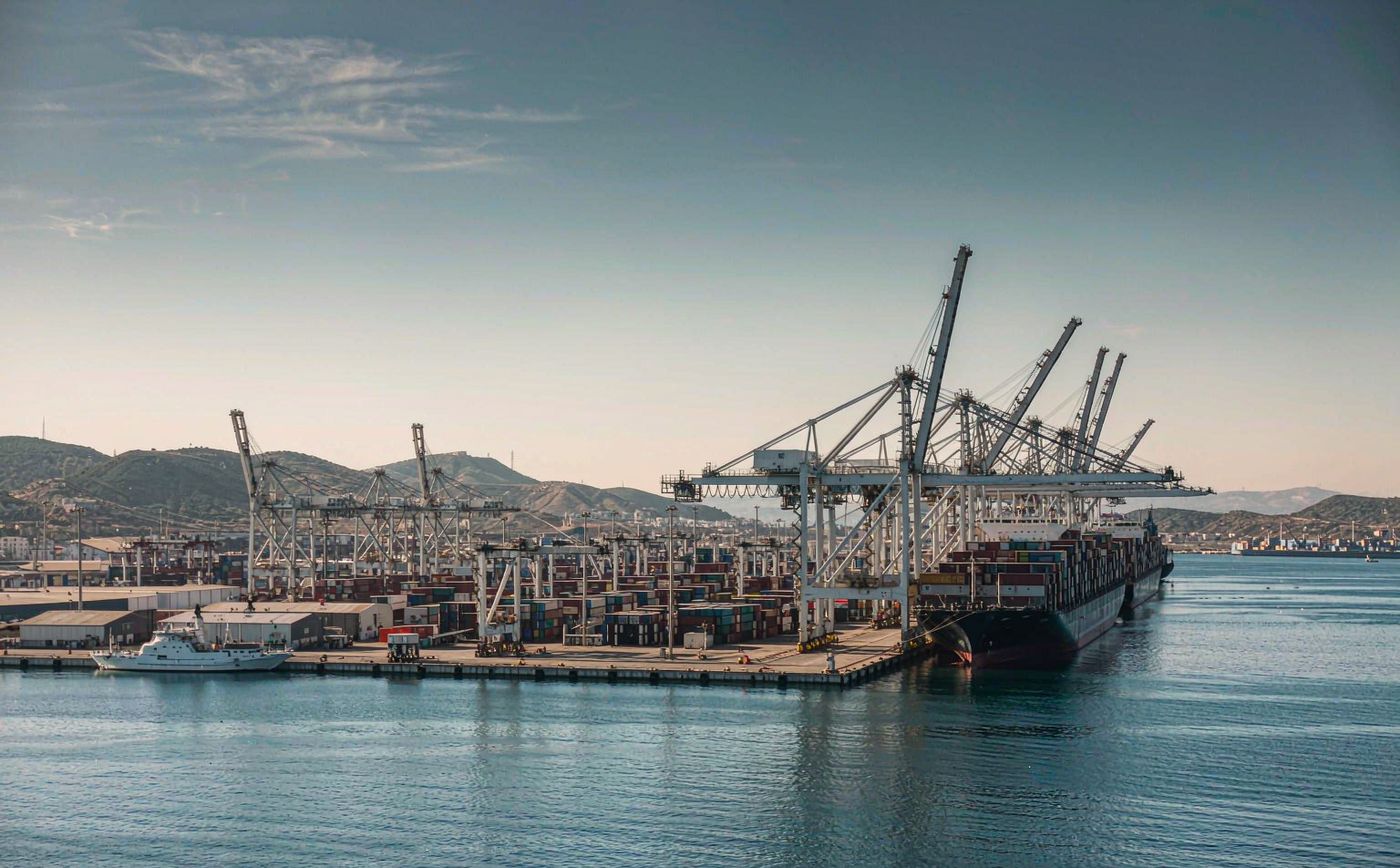 Mediterranean container port in bright daylight, tall ship-to-shore gantry cranes, rows of stacked shipping containers, a large container vessel berthed at the quay and low hazy hills beyond