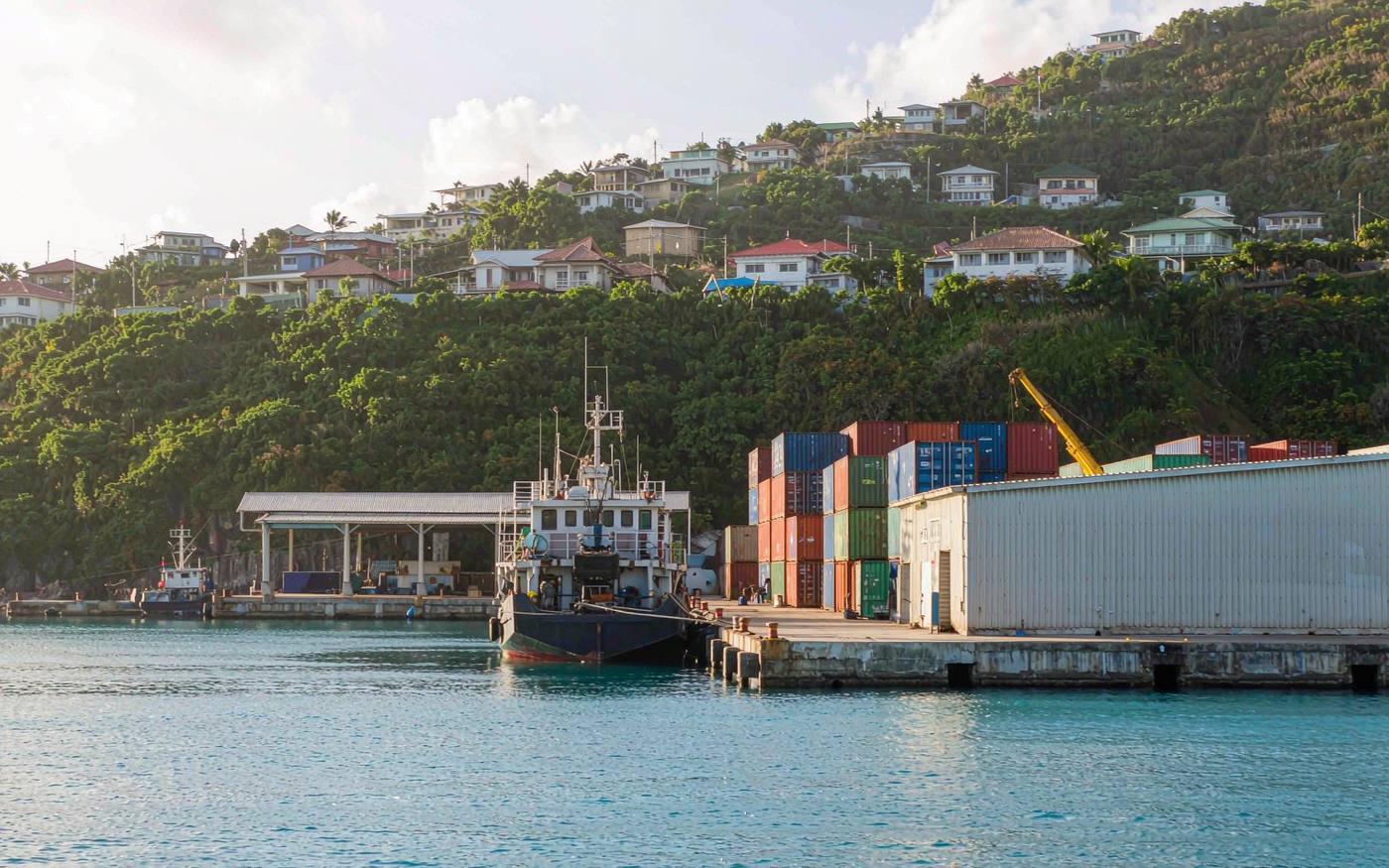 Tropical Caribbean harbour town with a small cargo berth, stacked containers, a cargo shed and green hillsides behind