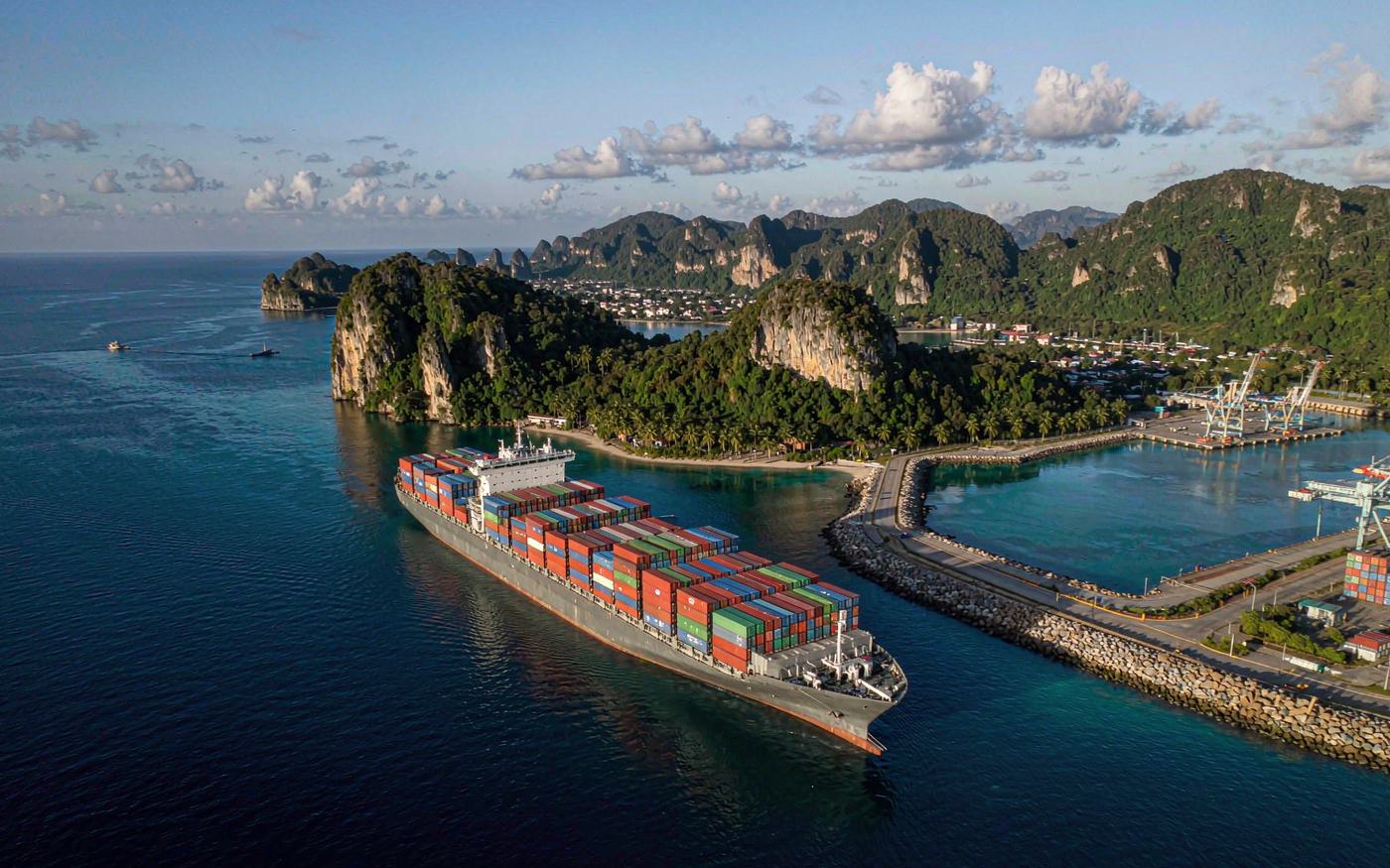 Aerial photograph of a container ship berthed at a deep-water Pacific island harbour, turquoise tropical water, green palm-covered limestone cliffs behind, a container yard with stacked boxes and gantry cranes, late afternoon light